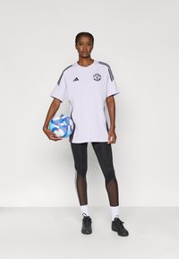 Nadměrná levandulová sportovní košile s černými akcenty a logem, doplněná černými legínami s síťovanými panely a bílými pruhy po stranách. Drží modrý fotbalový míč.