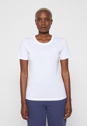 Vrouw met kort blond haar die een eenvoudig wit T-shirt met korte mouwen en gestructureerde blauwe broek draagt, staand tegen een effen lichte achtergrond.