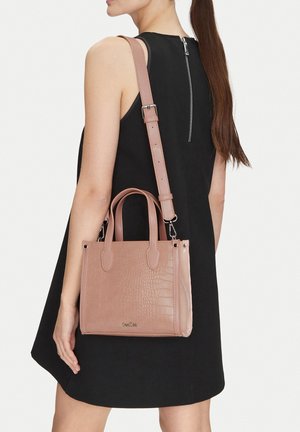 Žena v černých bezrukávových šatech se zadním zipem, nesoucí světle hnědou kabelku s krokodýlím vzorem, horními uchy a nastavitelným popruhem.