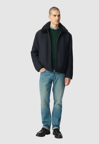 Hombre con una chaqueta negra de cuello de borrego, suéter verde, jeans azules y zapatos negros, de pie con una mano en el bolsillo contra un fondo liso.