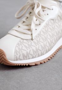 Beige sneaker med en tekstureret stofkrop og læderdetaljer. Har en hvid sål med et zigzag-mønster og flade snørebånd til fastgørelse.