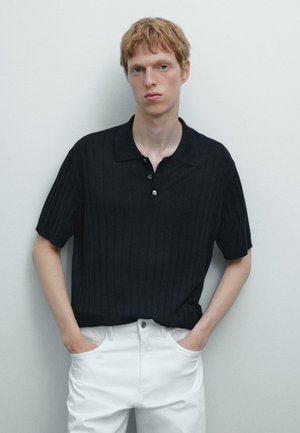 Sort ribbet polo skjorte med krave, tre knapper, korte ærmer, parret med hvide bukser, vist mod en neutral baggrund.