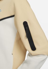 Manche de veste beige et blanche avec une poche verticale noire zippée sur le haut du bras et une bordure noire le long de la poitrine.