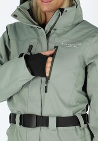 Light green waterproof jacket with a zipper, adjustable velcro cuffs, and a black belt. Features logo and multiple zippered pockets.