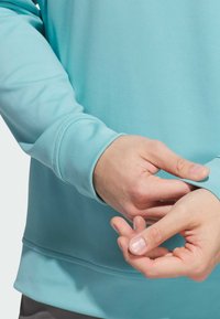 Světle modrý pulovr s dlouhými rukávy. Hladká látka, přiléhavé manžety a jemná textura. Viditelné ruce upravující manžetu rukávu.