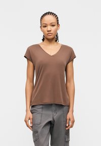 Νεαρό άτομο με πλεκτά μαλλιά, φορώντας καφέ μπλούζα με V λαιμόκοψη και κοντά μανίκια και γκρι παντελόνια cargo, στέκεται μπροστά σε απλό φόντο.