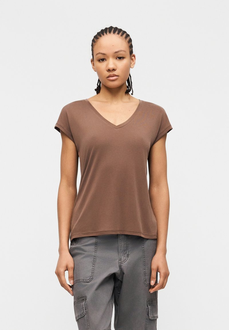 Νεαρό άτομο με πλεκτά μαλλιά, φορώντας καφέ μπλούζα με V λαιμόκοψη και κοντά μανίκια και γκρι παντελόνια cargo, στέκεται μπροστά σε απλό φόντο.