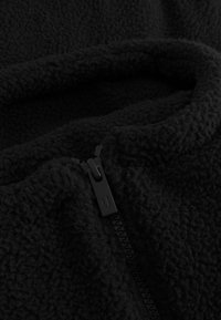 Tessuto in pile nero con una superficie morbida e texture. Presenta una cerniera nera che si avvolge lateralmente lungo la cucitura.