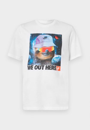 Camiseta de algodón blanca con un gráfico de un animal con un sombrero de cubo y gafas de sol frente a un fondo espacial. El texto dice "AQUÍ ESTAMOS".