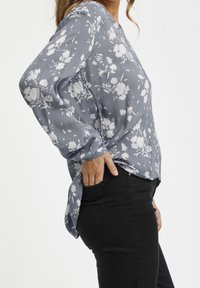 Blusa leggera a maniche lunghe grigia con motivo floreale bianco. Caratterizzata da una vestibilità morbida, scollo a V e tessuto texturizzato. Abbinata a jeans neri.