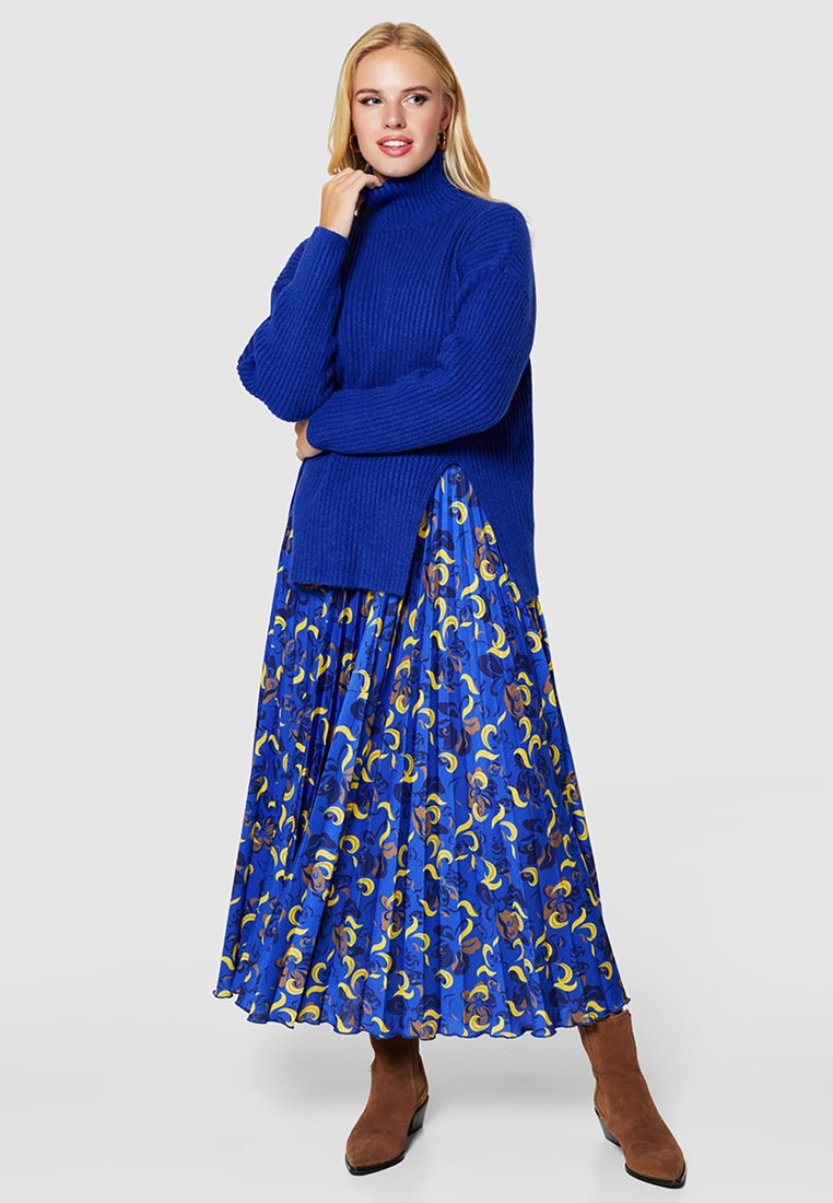 Blondynka w niebieskim swetrze z golfem, kwiecistej niebieskiej spódnicy plisowanej z żółtymi akcentami i brązowych botkach za kostkę, stojąca z ręką blisko twarzy.