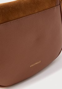 Bruine leren en suède handtassectie met zichtbare stiksels en goudkleurig COCCINELLE-logo op witte achtergrond.