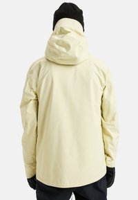 Gelbe wasserdichte Jacke mit Kapuze und elastischen Bündchen, von hinten angezeigt, kombiniert mit schwarzen Handschuhen und Hosen. Glatte, matte Textur.