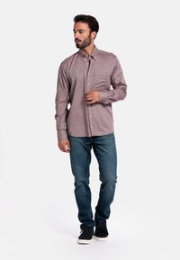 Langarmshirt in bordeaux met een subtiel patroon, kraag met knopen, en borstzak, gecombineerd met blauwe jeans en zwarte schoenen.