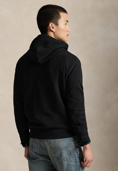 Man in black hoodie and blue jeans standing turned slightly away, looking to the right against a plain beige background.
