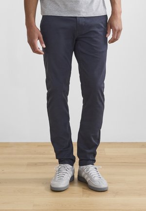 Man draagt slanke donkerblauwe broek en witte veterschoenen, staat op houten vloer tegen een grijze muur.