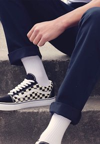 Sort og hvid ternede lærreds sneakers med hvide snørebånd, parret med hvide sokker, liggende på foldede marineblå bukser, der hviler på en stenoverflade.