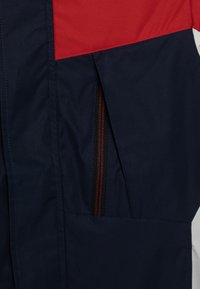 Tummansininen takki, jossa on punavalkoinen yläpaneeli, varustettuna vetoketjullisella taskulla punaisilla yksityiskohdilla. Sileä, teksturoitu kangas.