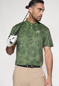 Žalia raštuota polo marškinėliai su tekstūruotu dizainu, trijų mygtukų apykakle ir išsiuvinėta logotipu, derinami su smėlio spalvos kelnėmis ir golfo lazda.