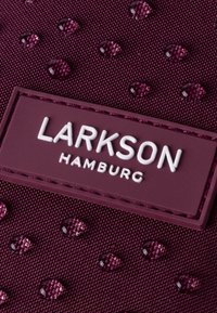 Tessuto bordeaux con una superficie testurizzata, caratterizzato da piccole gocce in rilievo e una toppa rettangolare bianca con la scritta "LARKSON HAMBURG".