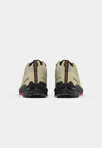Zapatos de exterior beige con una parte superior de tela texturizada, suela de goma negra y detalles rojos. Presentan una asa con un patrón a rayas.