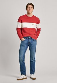 Sudadera a rayas rojas y blancas con logo, combinada con jeans azules y zapatillas blancas con suelas marrones. Conjunto casual presentado.