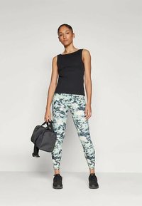 Svart ärmlös topp med åtsittande design. Mönstrade leggings i olika nyanser av grönt och grått. Modellen håller en svart sportväska och bär svarta sneakers.