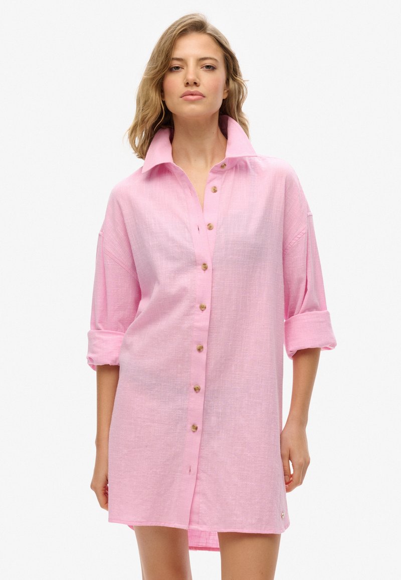 Vestido de camisa de lino rosa con un frente abotonado, gran cuello, mangas enrolladas y un pequeño detalle de logo en el dobladillo. Acabado de tela texturizada.