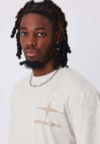 T-shirt beige in cotone con una texture logora. La parte anteriore presenta un logo sbiadito in marrone chiaro, completato da una collana in argento.