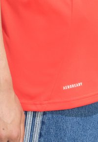 Rode Aeroready sporttop, met een zachte textuur, slank ontwerp en een wit logo aan de onderkant. Gecombineerd met blauwe denim.