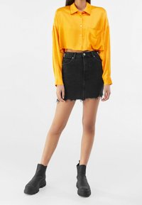 Camicia corta in satin arancione con chiusura a bottoni, abbinata a una minigonna in denim nero con orlo sfrangiato. Indossata con stivaletti neri alla caviglia.