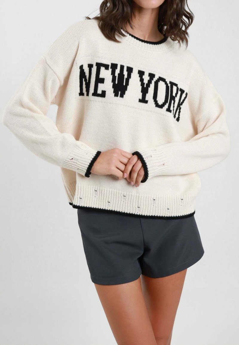 Maglione lavorato a maglia color crema con la scritta "NEW YORK" in lettere nere, dettagli neri su polsini e orlo, abbinato a pantaloncini scuri.