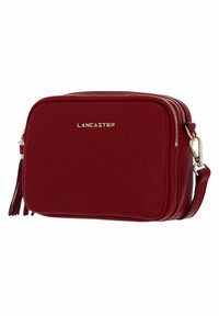 Borsa a tracolla in pelle rossa con forma rettangolare, dettagli in metallo dorato, doppie chiusure con zip e un elegante pon-pon su un lato.