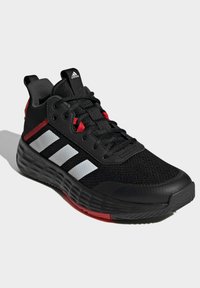 Zapatilla deportiva negra con parte superior de malla, tres franjas blancas, acentos rojos y suela texturizada. Presenta un diseño de cordones y un collar acolchado.