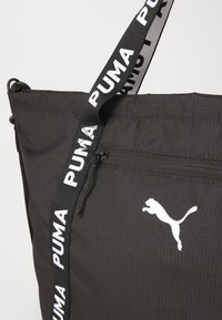 Svart tote-väska med dragkedja och en vit Puma-logotyp, med en märkesrem med "PUMA"-text. Enkla, stilren design för vardagsanvändning.