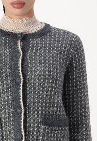 Pelēks adīts kardigans ar teksturētu rakstu, krēmīgas krāsas apdari un pogām. Ir kabata kreisajā pusē. Mīksts vilnas maisījuma materiāls.