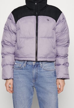 Bijgesneden lila en zwarte Calvin Klein gewatteerde jas met rits aan de voorkant, gecombineerd met lichtblauwe jeans.