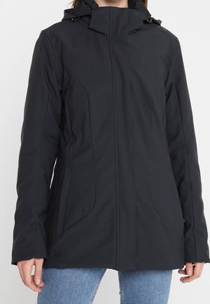 Kvinde iført sort hættejakke med lynlås foran og lommer, kombineret med blå jeans, står mod en ensfarvet baggrund.