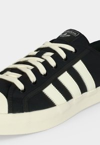 Άσπρο-μαύρο αθλητικό παπούτσι Adidas με λευκές κορδόνια, τρεις λευκές ρίγες στο πλάι και λευκή λαστιχένια σόλα.