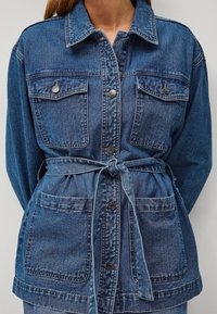 Giacca di denim in diverse tonalità di blu, con due tasche frontali, vita con cintura e colletto con bottoni metallici.