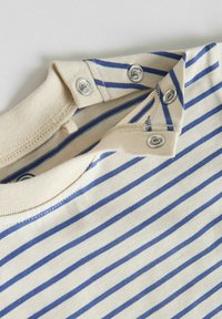 Encolure d'une chemise crème à rayures horizontales bleues, avec trois boutons-pression argentés partiellement ouverts au niveau de l'épaule.