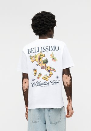 Άτομο με λεύκη στέκεται με την πλάτη, φορώντας λευκό μπλουζάκι με πολύχρωμο χάρτη της Ιταλίας και το κείμενο «BELLISSIMO Vacation Club».