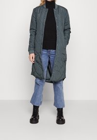 Lång, vadderad grön kappa med ståkrage, tryckknappar och sidofickor, matchad med en mörk tröja och ljusblå jeans samt svarta stövlar.