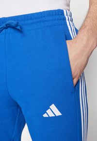 Blue athletic pants made of cotton blend, featuring white stripes along the sides and an embroidered white logo on the left thigh.