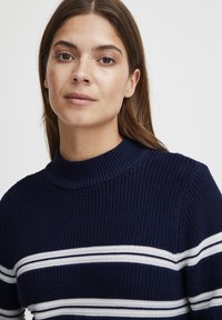 Marineblaues Strickpullover mit hohem Rippkragen, der horizontale weiße und hellblaue Streifen über die Brust und Ärmel aufweist.
