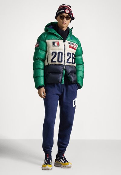 Steppjacke in Grün, Blau und Creme mit amerikanischen Flaggen und '2026' auf der Vorderseite; kombiniert mit marineblauen Jogginghosen und mehrfarbigen Sneakers.