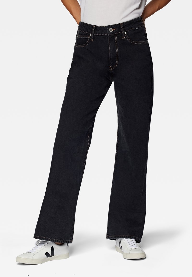 Personne portant un jean noir droit, des baskets blanches avec un logo "V" noir, debout devant un fond uni les mains sur les hanches.