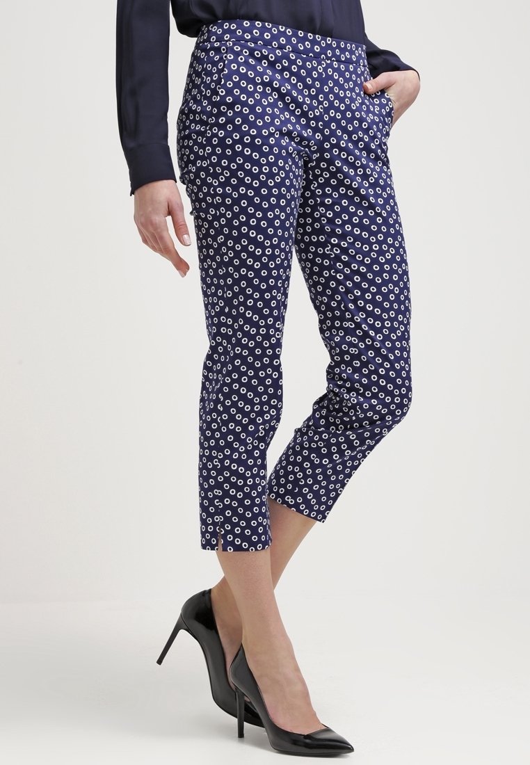 Pantalons navy raccourcis avec des motifs circulaires blancs, dotés de poches latérales et d'une texture lisse, associés à des chaussures à talons hauts noires.