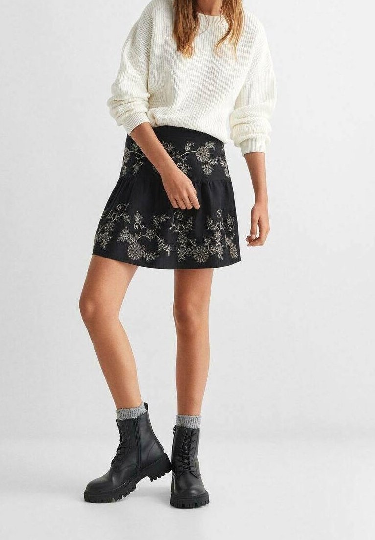 Femme portant un pull blanc, une jupe noire brodée, des chaussettes grises et des bottes de combat noires à lacets, debout avec une jambe légèrement pliée.