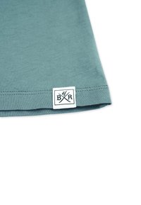 Türkisfarbiges Baumwoll-T-Shirt mit glatter Textur, versehen mit einem weißen Etikett, das am Saum mit einem schwarzen Logo-Design und gekreuzten Elementen gestickt ist.